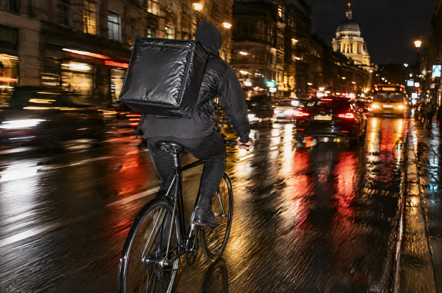 Un coursier à vélo, de dos, dans la rue la nuit